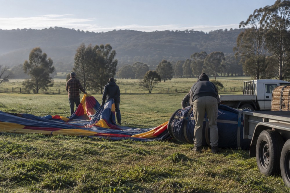 A balloon crew packing a deflated envelope into a large bag in an open paddock after landing