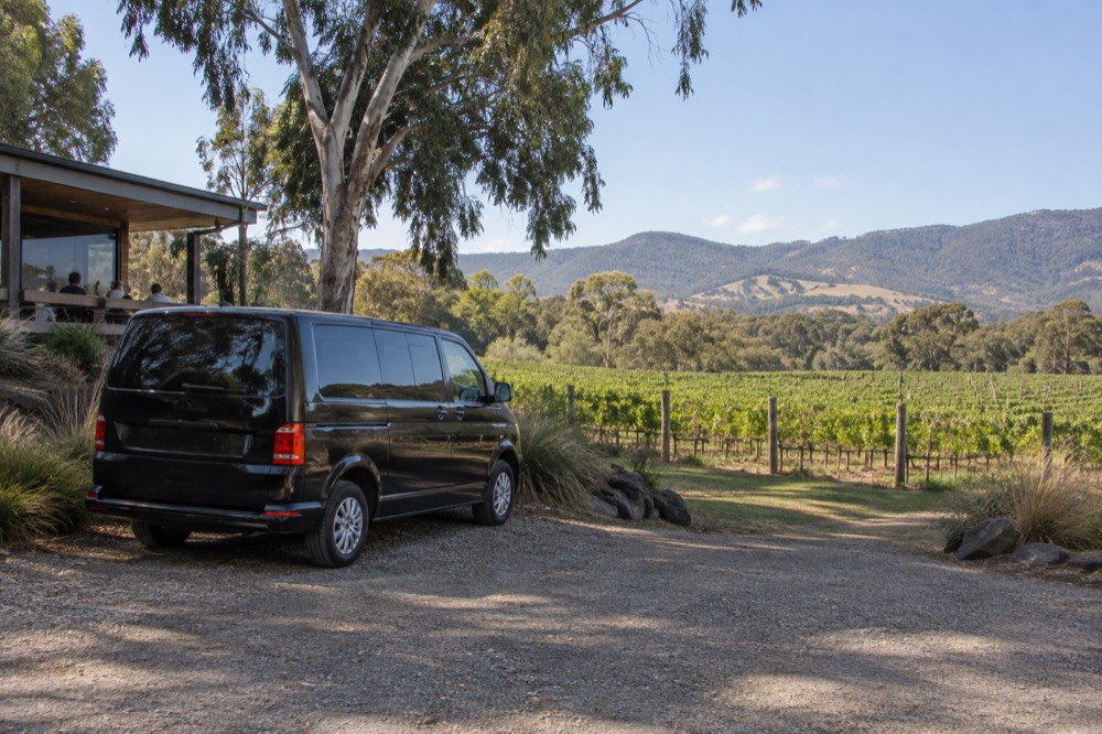 A small luxury van parked on a gravel driveway with a vineyard and hills behind it under a clear sky